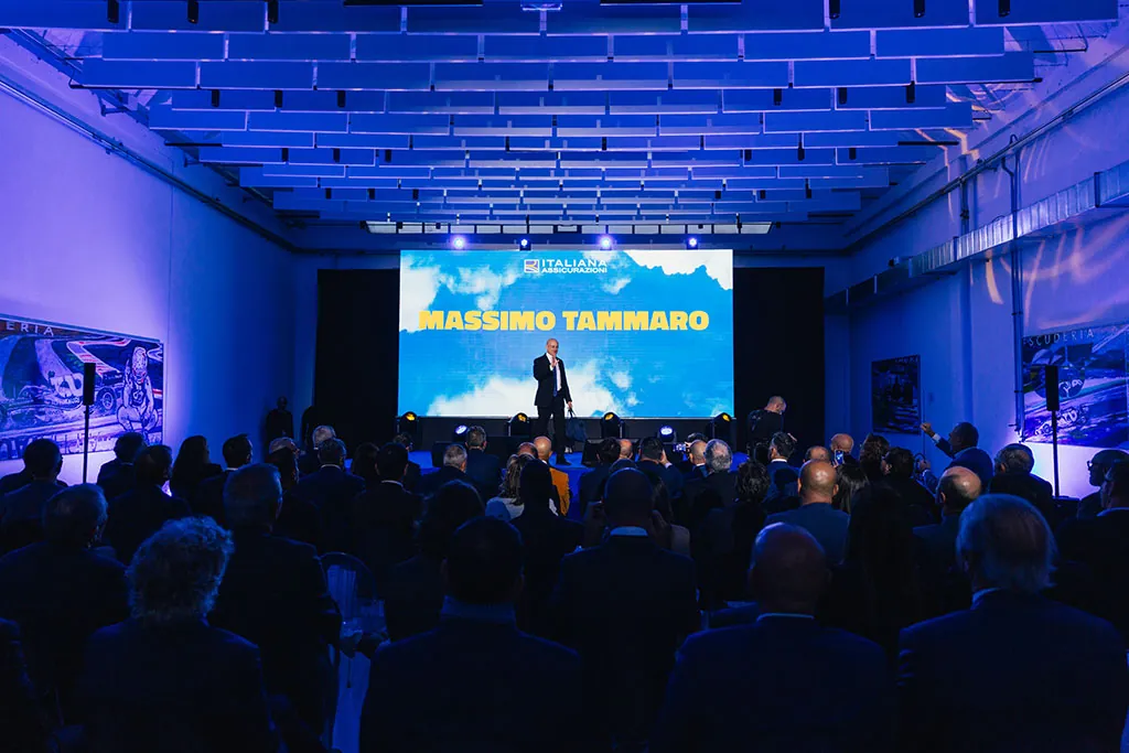Sala plenaria con luci blu, relatore sul palco e platea di partecipanti seduti
